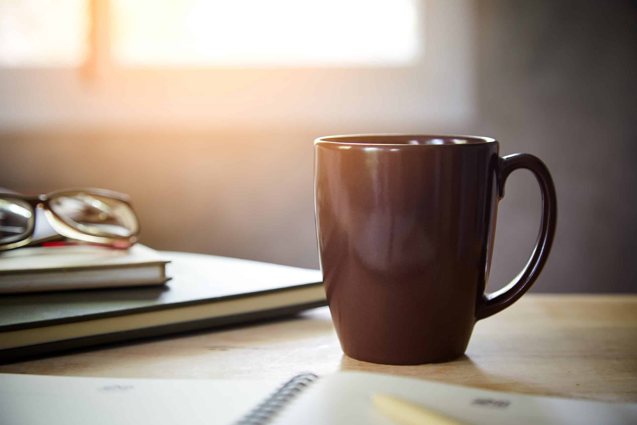 Morning coffee on the desk with sunlight from window, 
Coffee mug on the table with the blurred of notebook and glasses